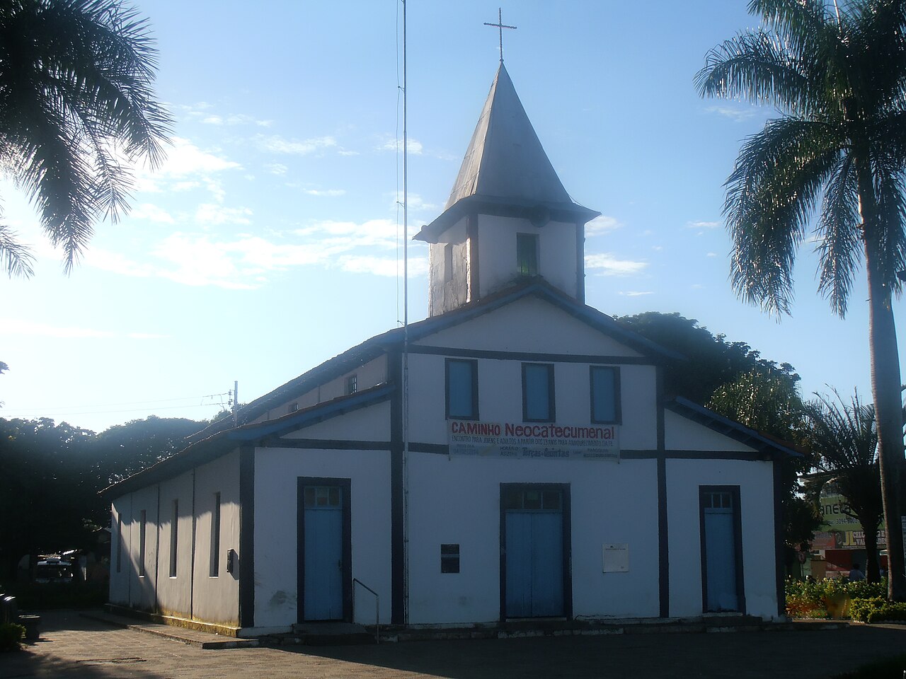 Paróquia Nossa Senhora Aparecida, igreja branca com torre pontiaguda e portas azuis, entre palmeiras na Praça da Matriz de Aparecida de Goiânia